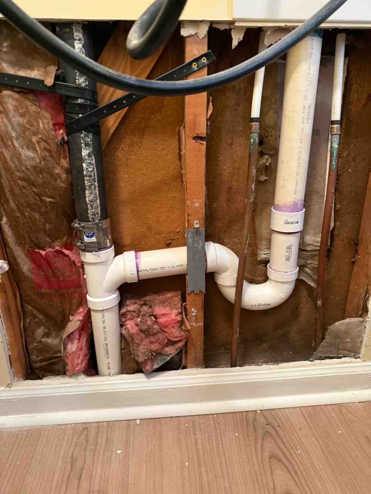 Exposed plumbing in a wall, including white PVC pipes, copper pipes, and black drainage pipes, with insulation and wood studs visible.