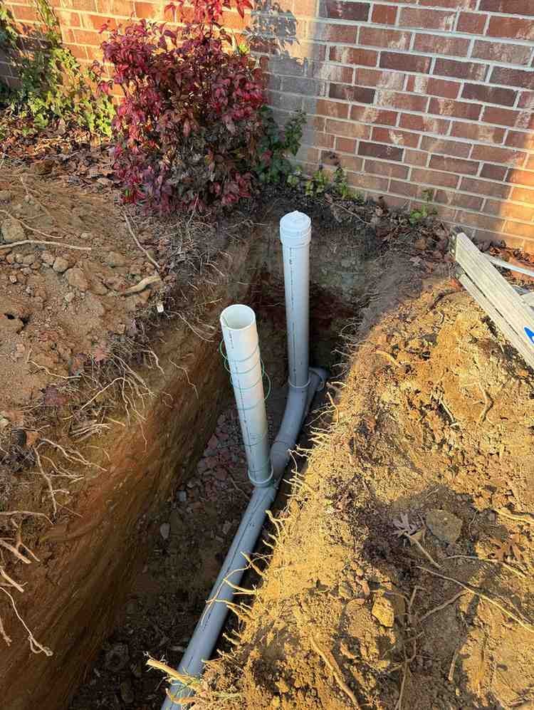 A trench dug next to a brick house foundation, showing plumbing pipes installed and two vertical white pipes.