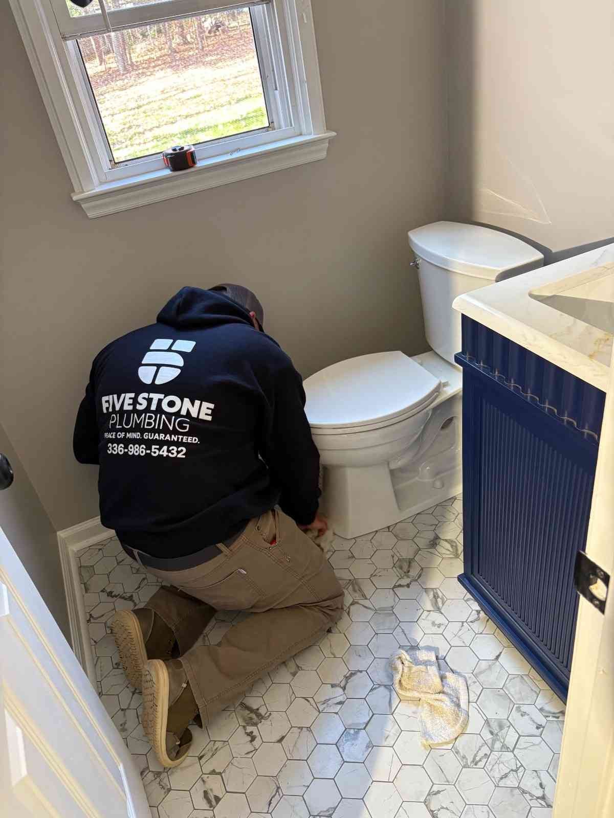 A plumber wearing a Five Stone Services hoodie kneels on hexagon tile, working on a toilet next to a blue vanity.