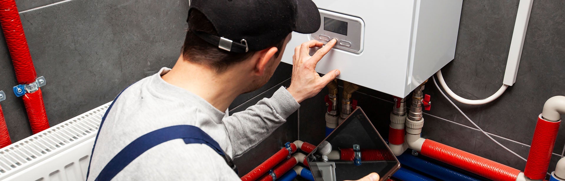 Hand of a plumber in a black cap adjusting the settings of a white boiler with red and blue pipes.